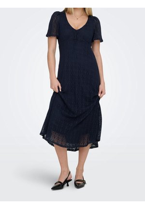 Kvinde iført en marineblå blonde midi-kjole med korte puffede ærmer, V-hals og sorte slingback sko med kitten hæl.