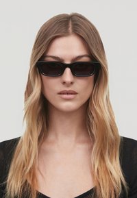 Occhiali da sole neri rettangolari con lenti scure, caratterizzati da una montatura spessa e una finitura opaca. Indossati da una persona con capelli lunghi e mossi.