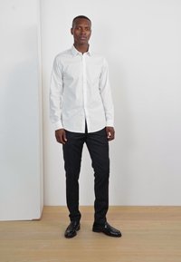Cămașă albă cu mânecă lungă și nasturi, pantaloni slim-fit negri, pantofi eleganți negri, stând pe un podea din lemn, în fața unui perete alb simplu.
