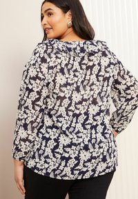 Mörkblå blus med blommönster i vitt, med en volangkrage och långa ärmar. Tillverkad av ett lätt, genomskinligt tyg.