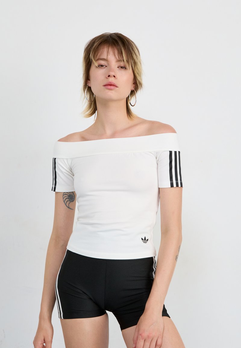 Balta off-the-shoulder palaidinė su juodais trimis juostelėmis ant rankovių, derinama su juodomis šortais su panašiomis juostelėmis. Rodoma Adidas logotipas.