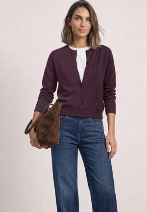Donna che indossa un cardigan viola con bottoni sopra una camicia bianca e jeans blu, che tiene una borsa marrone in camoscio su uno sfondo chiaro.