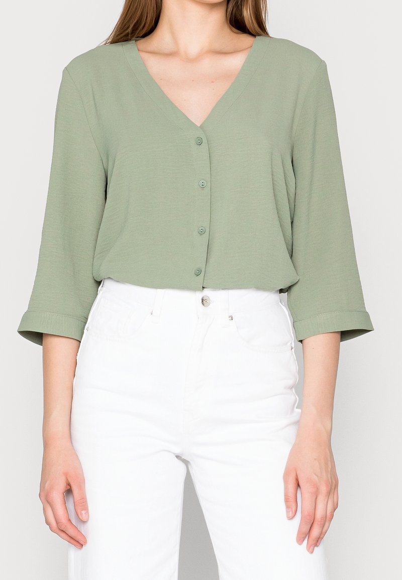 Femme portant un chemisier boutonné vert sauge à col en V avec des manches trois-quarts, associé à un pantalon blanc taille haute.