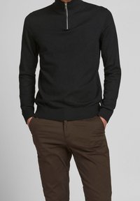 Maglione nero a maglia con collo alto e chiusura con zip corta, dotato di maniche lunghe e orlo a coste. I pantaloni marroni completano l'outfit.