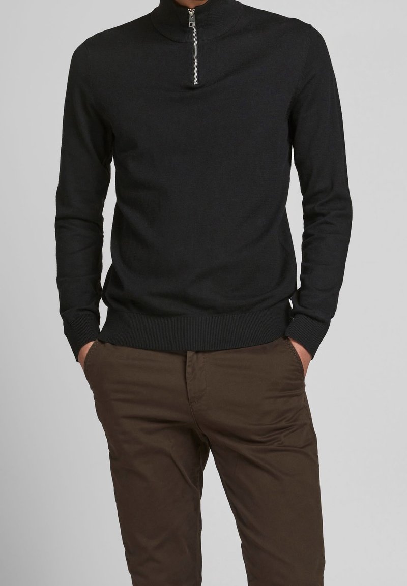Maglione nero a maglia con collo alto e chiusura con zip corta, dotato di maniche lunghe e orlo a coste. I pantaloni marroni completano l'outfit.