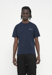 Μπλούζα από βαμβάκι σε ναυτικό μπλε χρώμα με λαιμόκοψη τύπου crew, κοντά μανίκια και μικρό λευκό λογότυπο στο αριστερό στήθος, συνδυασμένη με μαύρο παντελόνι cargo.