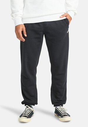 Pantalones de chándal negros con puños elásticos, bolsillos laterales y un logotipo en el muslo izquierdo. Están hechos de una tela suave con una textura lisa.