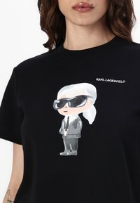 Juodas medvilnės marškinėliai su piešiniu, vaizduojančiu animacinį personažą kostiumu, akiniams nuo saulės ir baltais plaukais, su užsiuvinėtu tekstu "KARL LAGERFELD" viršuje.