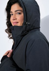 Černá kapucňová bunda s hladkým, matným povrchem. Obsahuje nastavitelné příslušenství, vysoký límec a přiléhavý střih.