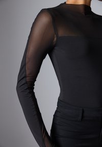 Fekete bodysuit hosszú, áttetsző hálós ujjal és sima, testre simuló fazonnal. Az anyag rugalmas, magas nyakú kialakítással és láthatatlan záródással rendelkezik.