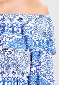 Abito off-the-shoulder in tessuto bianco con motivi floreali e geometrici blu. Presenta volant a strati e una vita elasticizzata.