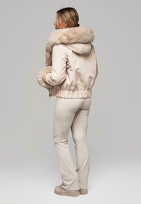Jasnoróżowa kurtka bomberka z kapturem i wykończeniem z futra ekologicznego; posiada ściągacz na dole i przy mankietach, w zestawie z dopasowanymi kremowymi spodniami i beżowym obuwiem.
