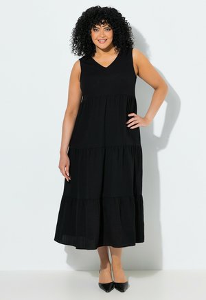 Uśmiechnięta kobieta z kręconymi włosami, ubrana w czarną bezrękawnikową sukienkę midi z falbanami i czarne buty na szpic, stojąca z jedną ręką na biodrze.