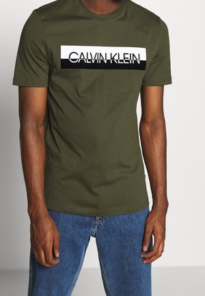 Muž nosící olivově zelené tričko Calvin Klein s černobílým logem, spárované s modrými džínami, stojící před jednoduchým pozadím.