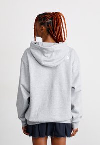 Personne avec de longs cheveux tressés portant un sweat à capuche gris clair et une jupe noire, de dos contre un fond blanc uni.