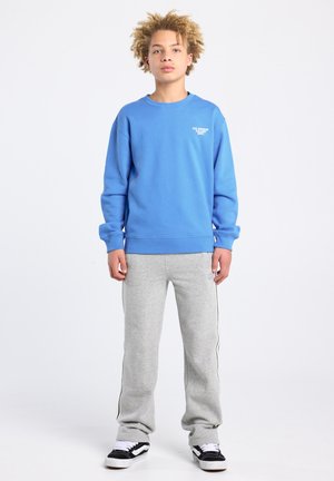 Blauwe trui met ronde hals, gecombineerd met grijze joggingbroek met zijstrepen. Model draagt zwarte sneakers met witte veters.