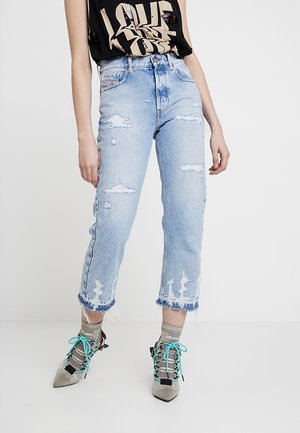 La personne porte un jean court bleu clair délavé, un haut sans manches noir à motif graphique, des chaussettes transparentes Diesel et des escarpins argentés pointus à lacets turquoise.