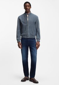 Giacca bomber in pelle grigia con chiusura a zip, polsini e colletto a costine; abbinata a jeans blu scuro e mocassini marroni.