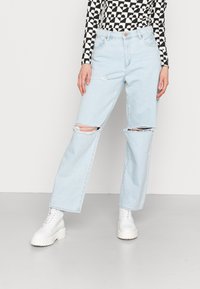 Jeans rasgados azul claro com um corte descontraído, apresentando áreas desgastadas nos joelhos, combinando com botas brancas de atacador chunky.