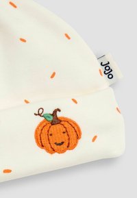 Weißes Baby-Mützchen mit orangefarbenem gesticktem Kürbisgesicht und kleinen verstreuten orangenen Strichen, mit einem "JoJo"-Textiletikett an der Seite.