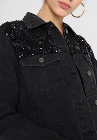 Mustat farkkutakki, jossa on paljettikoristeet harteilla, nappisuljenta, rintataskut ja rento istuvuus.