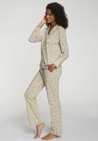 Frau mit lockigem Haar, die beige Pyjamas mit roten Zuckerstangenmustern trägt, steht barfuß da, eine Hand an das Gesicht gelegt, die andere in der Hosentasche.