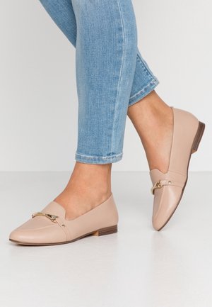 Fødder iført beige læder loafers med gulddetalje på spænde, parret med lyseblå skinny jeans mod en ensfarvet baggrund.