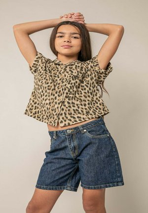 Fille aux longs cheveux portant une chemise à manches courtes imprimée léopard et un short en denim, posant les mains croisées derrière la tête.