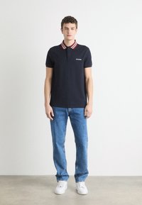 Ναυτικό πόλο μπλουζάκι με κόκκινο και άσπρο ριγέ γιακά, διαθέτει ένα μικρό λογότυπο και συνδυάζεται με ανοιχτό μπλε τζιν και λευκά αθλητικά παπούτσια.