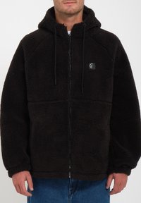 Veste en polaire noire avec capuche, fermeture éclair et cordons de serrage ajustables. Dotée d'un tissu doux et texturé ainsi que d'un logo sur la poitrine.