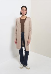 Beige lång cardigan i mjukt stickat material med framknappar, ihop med en brun topp och mörkblå jeans. Enkelt mönster, lätt textur.
