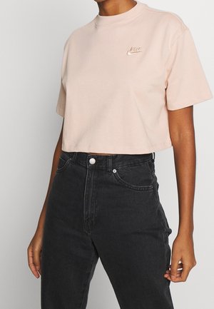 Persona con una camiseta corta rosa claro de Nike y jeans negros de cintura alta, de pie contra un fondo liso.