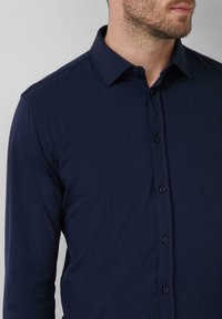 Chemise à manches longues bleu marine avec col à boutonnière, fabriquée en tissu lisse, dotée d'une patte de boutonnage et de coutures d'épaule.