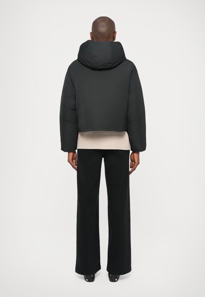Chaqueta acolchada negra recortada con capucha, combinada con un suéter texturizado de color beige claro y pantalones negros de pierna ancha. Acabado suave y mate.