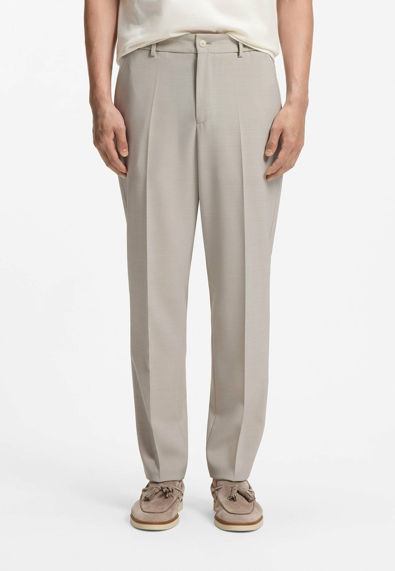 Homme portant un pantalon beige plissé associé à des mocassins en daim beige à glands et une chemise blanche rentrée.