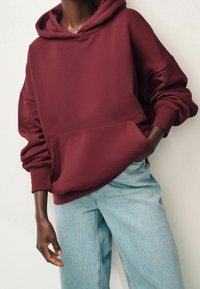 Personne portant un sweat à capuche ample couleur bordeaux avec poche kangourou et jean denim bleu clair, posant avec une main dans la poche devant un fond neutre.