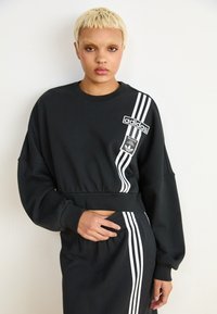 Melnā īsā džemperis ar garām piedurknēm, baltā Adidas logo un trim svītrām sānos, izgatavots no mīksta materiāla, ar brīvu piegriezumu.