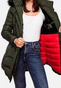 Personne portant un jean bleu foncé et un haut blanc, tenant ouverte une doudoune vert olive avec une doublure en fourrure rouge vif et noire.