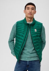 Grüne gepolsterte Weste mit hohem Kragen, mit horizontaler Steppung und einem kleinen Logo-Patch, getragen über einem strukturierten grünen Pullover.