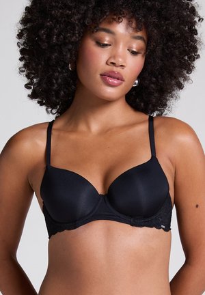 Vrouw met krullend haar die een zwarte gewatteerde beha met kantdetails draagt, kijkt naar beneden tegen een egale lichte achtergrond.