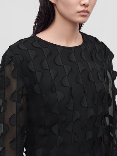 Gros plan sur une personne portant un chemisier noir à manches transparentes et aux motifs géométriques en relief.