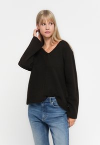 Svart genser med v-utringning, ribbede mansjetter og nederkant. Laget av mykt materiale, har den en avslappet passform og en subtil vertikal sømdetalj. Kombinert med blå jeans.