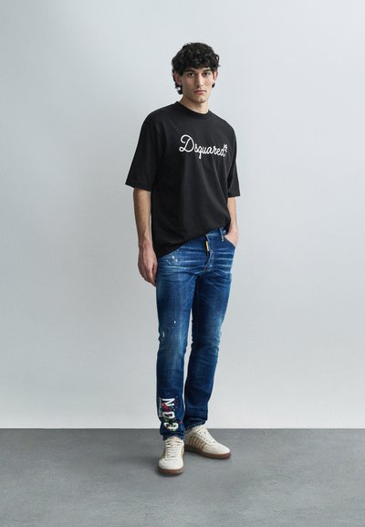 Black T-shirt with white "Dsquared2" logo, blue jeans with paint splatters, and beige sneakers. Casual fit with short sleeves.