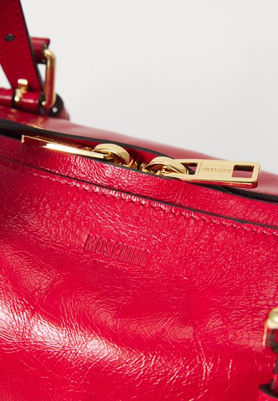 Sac à main en cuir rouge avec une surface texturée, des accessoires en or et un logo embossé sur le côté. Comprend une fermeture éclair et une bandoulière réglable.