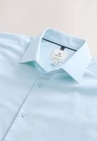 Hellblaues, mittelgroßes Hemd mit normaler Passform, spitzem Kragen und sichtbarem Markenlabel "NEXT" am Krageninneren.