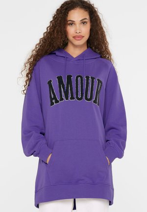 Woman with curly hair wearing a purple hoodie with black "AMOUR" lettering and white pants, hands in front pocket.