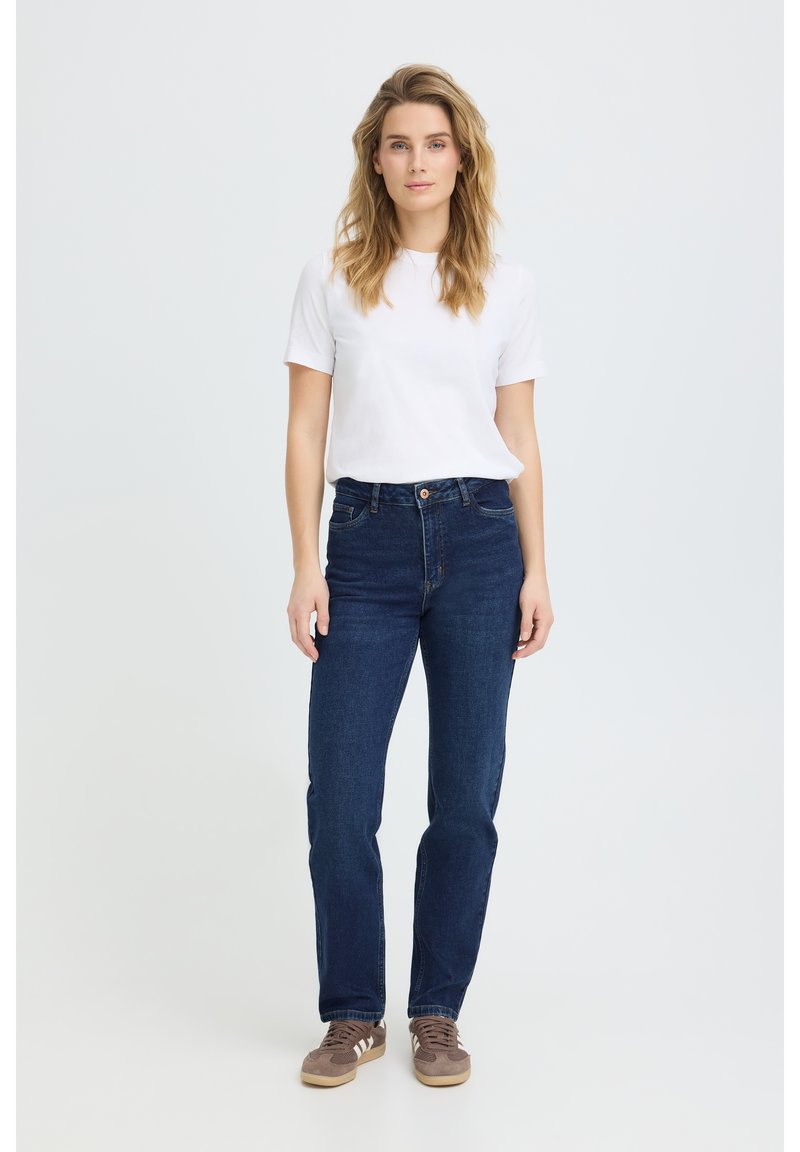 Kvinde med langt blondt hår iført en hvid T-shirt med korte ærmer, mørkeblå jeans og brune sneakers står foran en ensfarvet baggrund.