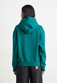 Felpa color teal realizzata in tessuto morbido, con un ampio cappuccio, polsini e orlo a coste. Indossata con pantaloni neri a righe sottili.