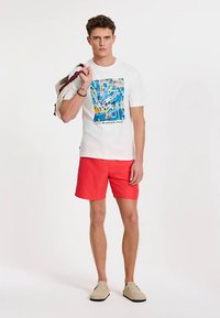 Weißes Baumwoll-T-Shirt mit farbenfrohem Grafikdesign, kombiniert mit knallroten Shorts und beigen Slip-On-Schuhen. Trägt eine Tasche über einer Schulter.
