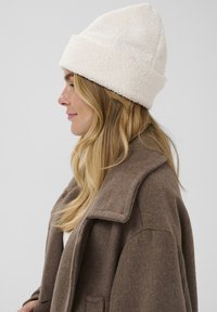 Biely, nadýchaný fleecový zimný čiapka s prehnutým okrajom, v kombinácii s hnedým, textúrovaným kabátom. Čiapka má uvoľnený, kužeľovitý tvar.